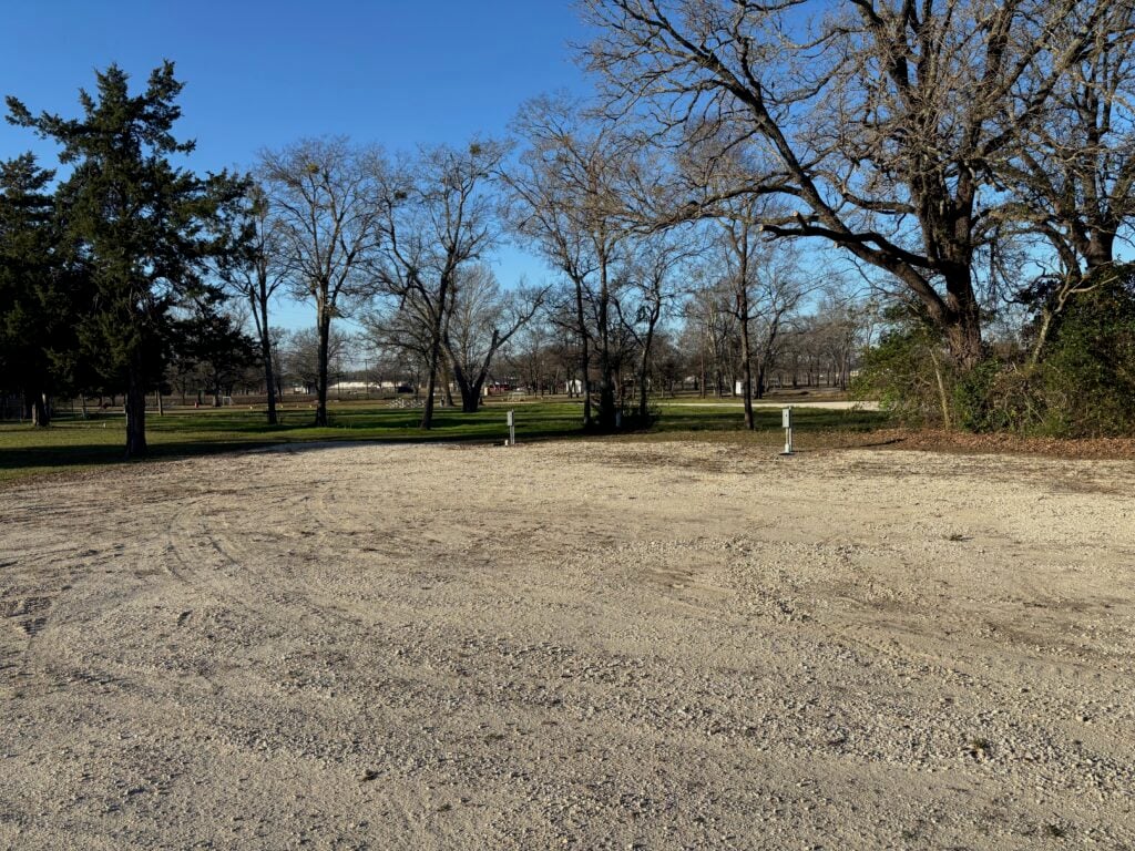 A gravel lot with electrical hookups, surrounded by leafless trees and grass under a clear blue sky.